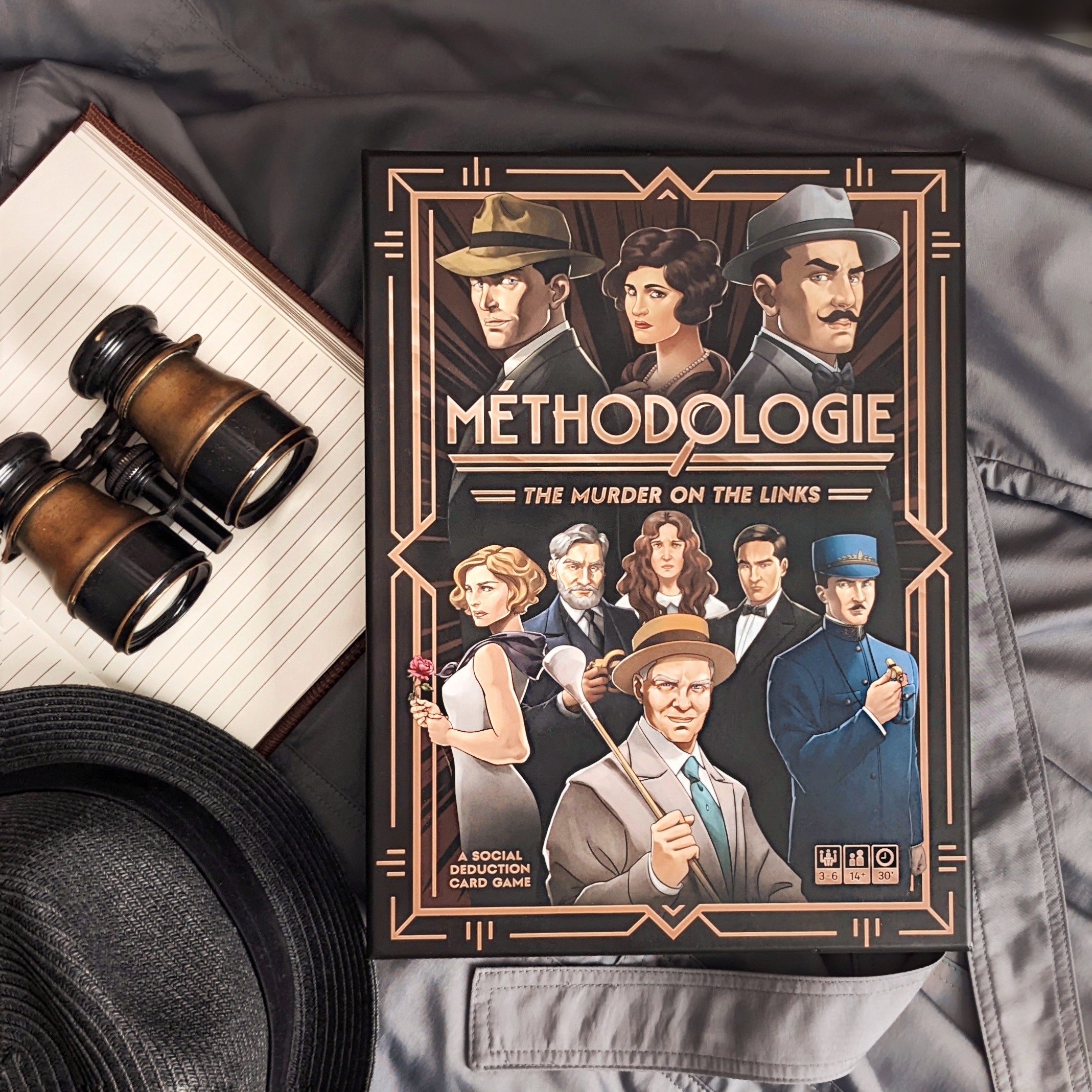 A photo of the board game box along with a notebook, pair of binoculars, and a hat - all on top of a trench coat.